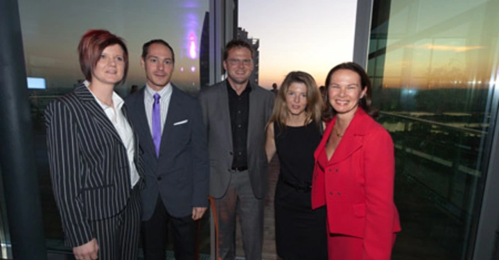 Galerie-Bild 54: Fünf Menschen stehen zusammen lächelnd in Geschäftsbekleidung vor verglasten Fenstern mit einem Sonnenuntergang und einem beleuchteten Gebäude im Hintergrund. Sie scheinen auf einer Veranstaltung zu sein.