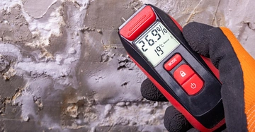 Hand mit Arbeiterhanschuhen hält ein Hygrometer zur Bestimmung der Feuchtigkeit an eine Steinmauer. Das Display zeigt 26,9 % Feuchtigkeit im Mauerwerk.