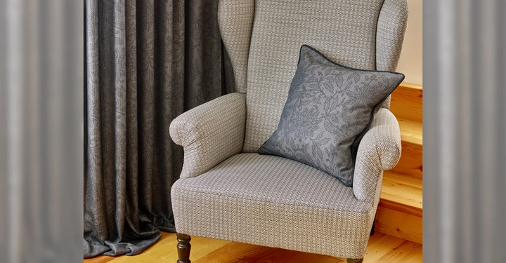 Galerie-Bild 5: Grau gepolsterter Sessel steht auf Holzfußboden mit einem dunklen dekorativen Kissen darauf, dahinter ein grauer Vorhang und eine Holztreppe.