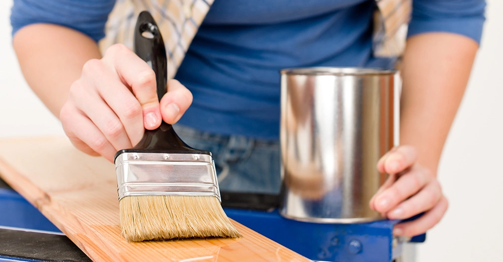 Eine Person trägt mit einem Pinsel lackähnliche Flüssigkeit auf ein Holzbrett auf wobei eine Metallfarbe-Dose auf einem Tisch daneben steht