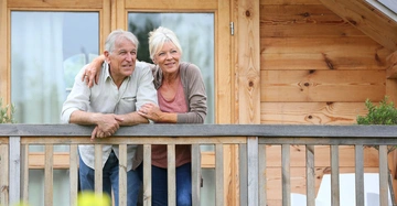 Älteres Paar lehnt auf dem Geländer einer Holzterrasse eines modernen Holzhauses lächelnd und entspannt.