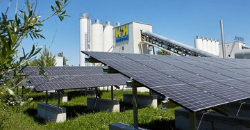 Solarmodule werden in der Nähe eines Betonwerks mit mehreren Silos betrieben umgeben von Grünflächen bei sonnigem Wetter. Text auf dem Gebäude: "K K BETON".