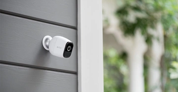 Weiße Überwachungskamera mit der Beschriftung arlo an einer grauen Hauswand montiert, Blick auf den grünen, verschwommenen Garten im Hintergrund.