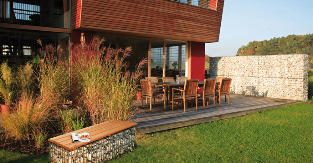 Gartentisch und Stühle stehen auf einer Holzterrasse vor einem modernen Gebäude aus Holz und rotem Putz umgeben von hohem Ziergras und einem Steinzaun im Hintergrund Rasen und bewaldete Hügel.