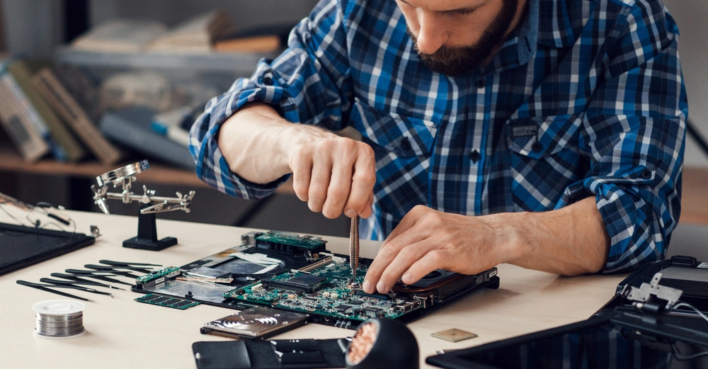 Person repariert eine Computerplatine auf einem Tisch mit Werkzeugen und Nadeln, umgeben von Büchern und Bürobedarf in einem Arbeitszimmer.