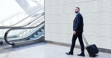 Ein Mann im Anzug zieht einen Rollkoffer hinter sich her in einem modernen, hellen Gebäude mit Rolltreppen im Hintergrund. Text: HYPO VORARLBERG.