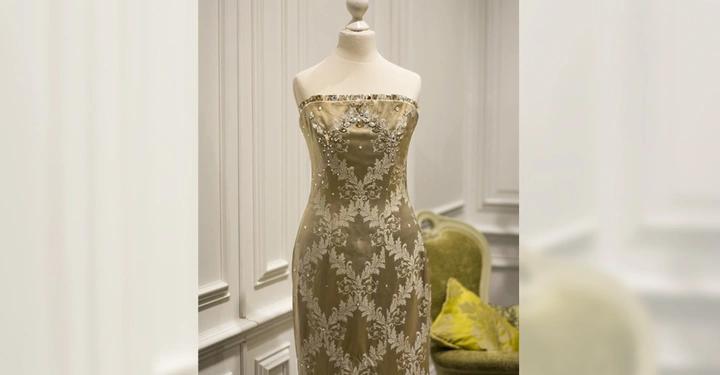 Galerie-Bild 4: Eine Schneiderpuppe trägt ein bodenlanges, trägerloses Kleid mit silberner Verzierung und Stickerei in einem eleganten Raum mit eine grünem Sessel und weiß getäfelten Wänden.