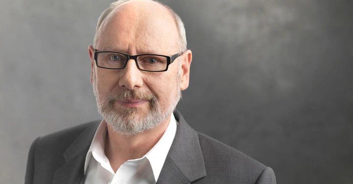 Ein Mann mit Brille und grauem Bart schaut ruhig in die Kamera trägt einen grauen Anzug und steht vor einem grauen Hintergrund