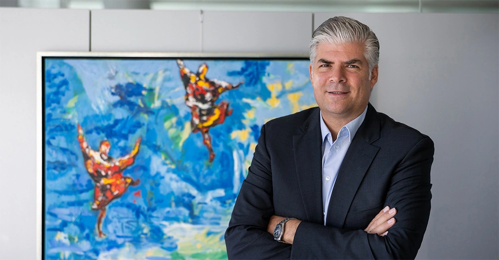 Geschäftsmann mittleren Alters in schwarzem Anzug und Krawatte steht mit verschränkten Armen vor hellem Bild mit orangen Clownfischen. 