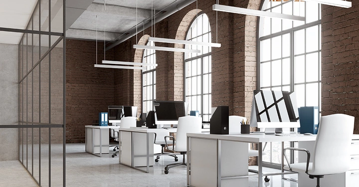 Weiße Bürostühle und Schreibtische mit Computern und Aktenordnern stehen in einem modernen Büro mit Backsteinwänden und großen Bogenfenstern vor einer verglasten Trennwand.