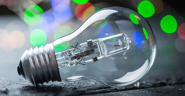 Eine Glühbirne liegt auf einer dunklen Oberfläche während im Hintergrund unscharfe bunte Lichter leuchten.