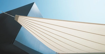Ein großes beige Sonnensegel erstreckt sich am Seil befestigt über einen modernen dunklen Gebäudeeingang vor einem klaren blauen Himmel.