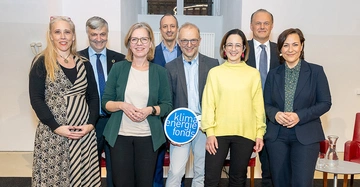 acht Personen (4 Männer, 4 Frauen), darunter Leonore Gewessler stehen auf einer kleinen Bühne, im Hintergrund ein Fenster, ein Mann im Vordergrund hält ein kleines Schild des Klilma- und Energiefonds in die Kamera