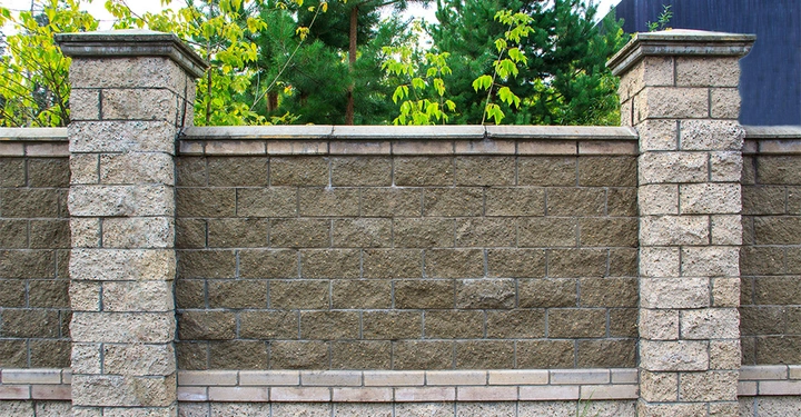Steinmauer steht still zwischen zwei Pfeilern umgeben von grünen Bäumen und blauen Himmel im Hintergrund.