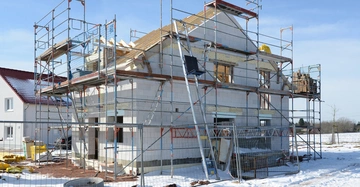 Ein Haus im Bau mit Gerüsten umgeben auf schneebedecktem Boden in einer Wohnsiedlung tagsüber bei klarem Himmel.