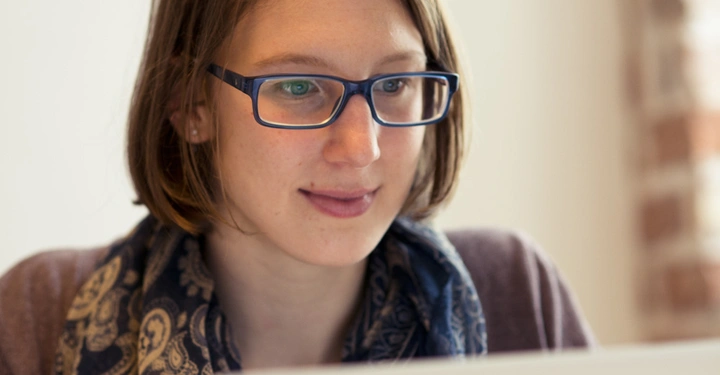 Galerie-Bild 12: Frau mit Brille arbeitet konzentriert an einem Laptop im Innenraum vor einer weißen Wand und einem teilweise sichtbaren Fenster mit ziegelrotem Rahmen.