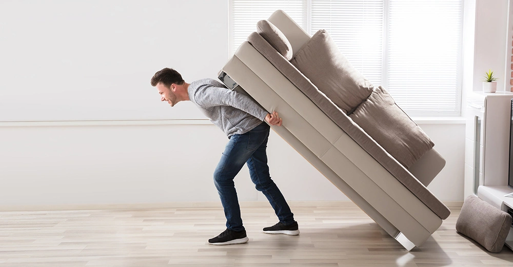 Mann mit gequältem Gesichtsausdruck schleppt ein Sofa auf dem Rücken durch eine leere Wohnung