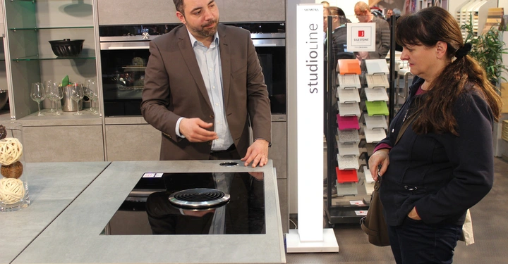 Galerie-Bild 3: Ein Mann zeigt einer Frau einen Herd in einem modernen Küchengeschäft mit Siemens StudioLine. Im Hintergrund sind Regal mit Gläsern und eine Farbmusterausstellung zu sehen.