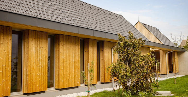 Galerie-Bild 2: Haus mit Holzwandverkleidung und dunkelgrauem Satteldach in einem grünen Garten mit Baum im Vordergrund an einem bewölkten Tag