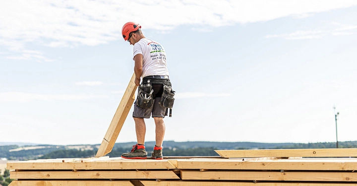 Galerie-Bild 4: Arbeiter hebt Holzbrett auf einer Baustelle am hellen Tag an trägt roten Helm Werkzeugtasche und T-Shirt mit dem Text Heinrich & Bock GmbH Altplya Alm Asplet ein hart PS δο Holz steht im Hintergrund.
