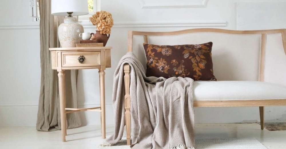 Helles Holzsofa mit weißem Polster, braunem Blumenkissen und beiger Decke. Daneben ein Beistelltisch mit dekorativer Lampe und Trockenblumen. Die Szene wirkt klassisch und gemütlich in neutralen Naturtönen vor weißem Hintergrund.