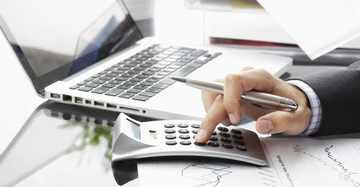 Eine Hand benutzt einen Taschenrechner auf einem Schreibtisch neben einem Laptop und einem Blatt Papier mit farbigen Diagrammen.