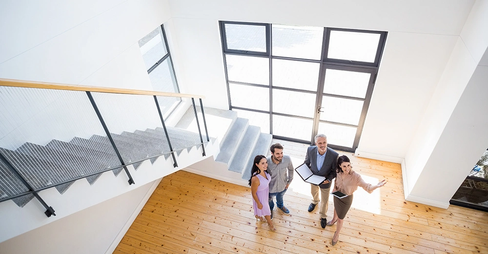 Vier Personen stehen und schauen nach oben in einem lichtdurchfluteten Raum mit Holzboden und großen Fenstern; eine Treppe mit Geländer führt nach oben.