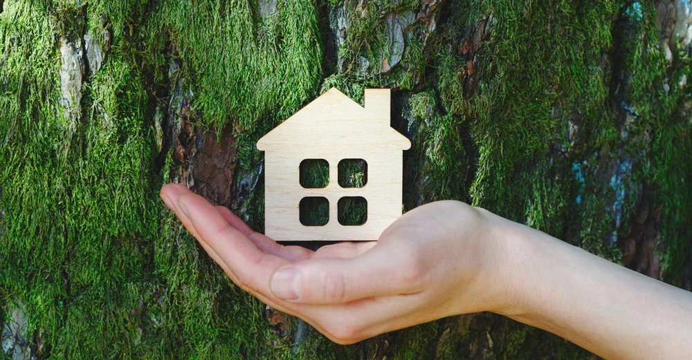 Hand hält kleines Holzhausmodell vor einem mit moos bewachsenem Baumstamm