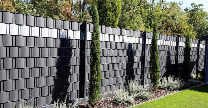 Hoher dunkelgrauer Zaun mit rechteckigen Mustern steht in einem Garten. Vor dem Zaun sind schlanke grüne Zypressen und niedrig wachsende Pflanzen auf einem Mulchbeet zu sehen. Bäume im Hintergrund.
