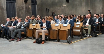 Erwachsene Menschen sitzen in einem alten Konferenzraum auf hölzernen Stühlen und halten Notizen oder elektronische Geräte in den Händen während einer Veranstaltung.