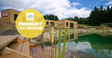 Holzgebäude liegend Graspflanzen neben einem Teich grünem Wasser auf einem Hügel vor einem bewaldeten Hintergrund blauer Himmel und Wolken mit einem gelben Kreis und folgendem Text Kalender 49 PRODUKT DER WOCHE.