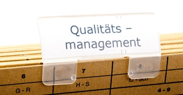 Ein Registerkartenetikett zeigt den Text Qualitäts-Management auf mehreren braunen Ordnern in einer Ablage vor einem weißen Hintergrund.