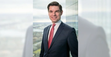 Ein Mann in einem dunkelblauen Anzug mit roter Krawatte lächelt in die Kamera. Im Hintergrund sind Hochhäuser und eine Stadtlandschaft durch ein Glasfenster zu sehen.