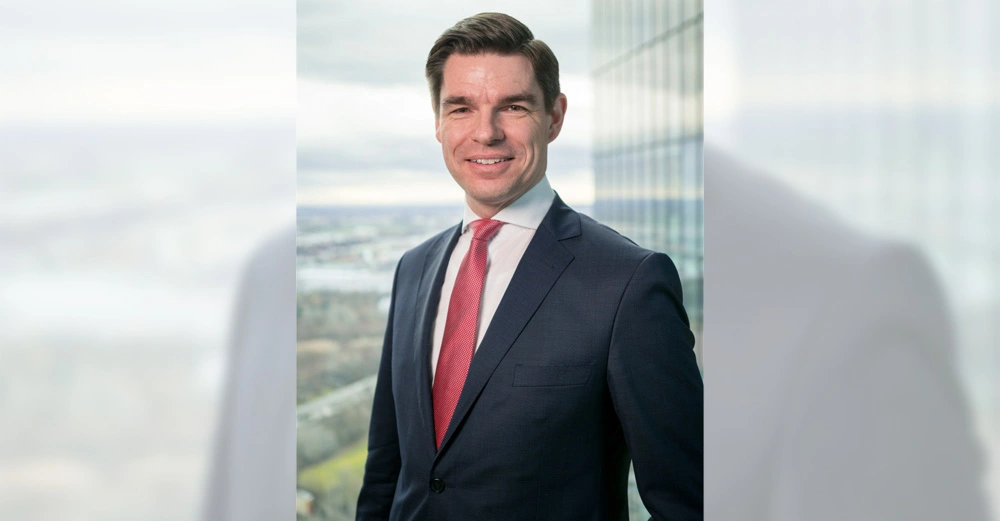 Ein Mann in einem dunkelblauen Anzug mit roter Krawatte lächelt in die Kamera. Im Hintergrund sind Hochhäuser und eine Stadtlandschaft durch ein Glasfenster zu sehen.