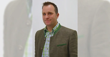 Mann posiert lächelnd in braunem Trachtenjanker und kariertem Hemd in einem hellen Raum