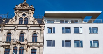 Ein altes Gebäude mit verzierter Fassade und Balkonen steht neben einem modernen weißen Gebäude mit rechteckigen Fenstern unter einem klaren blauen Himmel.