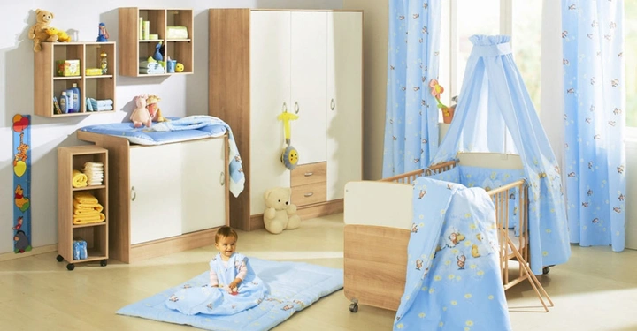 Galerie-Bild 6: Kleinkind sitzt auf einer blauen Spieldecke in einem hellen Kinderzimmer mit Holz- und weißen Möbeln im Hintergrund. Ein Babybett, Kommode, Schrank und Regale sind umgeben von hellblauen Vorhängen.