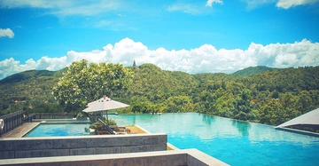 Großer Infinity-Pool an einem sonnigen Tag mit Liegestühlen und Sonnenschirm daneben Hügelige grüne Landschaft und blauer Himmel im Hintergrund.