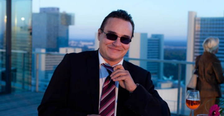 Galerie-Bild 17: Mann trägt Anzug und Sonnenbrille bindet seine Krawatte Abendveranstaltung auf einer Dachterrasse mit städtischer Skyline im Hintergrund rechter Bildrand Tisch mit Gläsern und Blume.