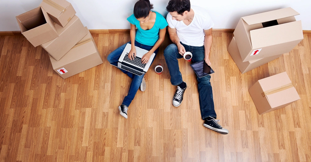 Frisch eingezogen: Pärchen von oben sitzt mit Kaffeetassen in der Hand, auf poliertem Holzlaminatboden in leerem Raum, Umzugskartons rundherum