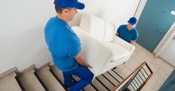 Zwei Männer tragen ein weißes Sofa eine Treppe hinauf in einem Treppenhaus mit weiß gestrichenen Wänden und blauer Tür.