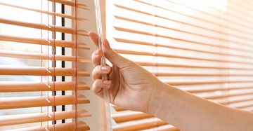 Frau dreht an Bedienstange von Innenjalousien aus Holz, noch sind die Innenjalousien offen, schönes, weiches, angenehmes Licht