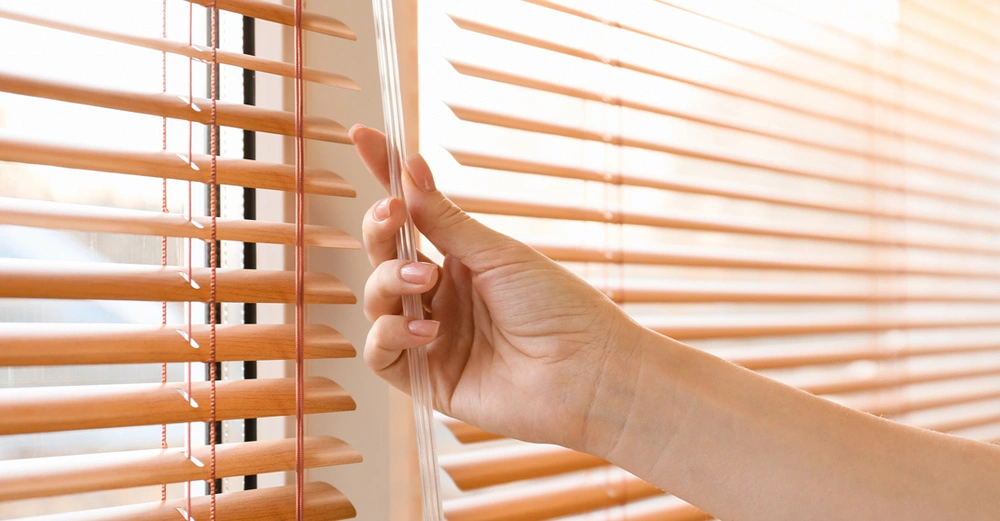 Frau dreht an Bedienstange von Innenjalousien aus Holz, noch sind die Innenjalousien offen, schönes, weiches, angenehmes Licht