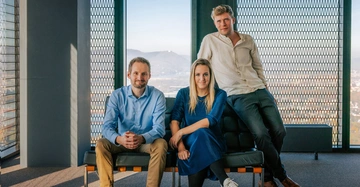 Drei Personen sind in einem modernen Raum mit Blick auf die Berge. Zwei Personen sitzen auf einem schwarzen Sofa, während eine Person auf der Sofalehne steht.