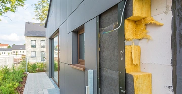 Hausisolierung wird erneuert neben einem Gartenweg in einer Wohngegend mit mehrstöckigen Häusern im Hintergrund gelber Dämmstoff freigelegt mit aufgebrachten Isolierplatten die Isolierung zeigen. Text: 1025 X 36