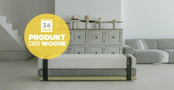 Ein graues Sofa steht in einem minimalistisch eingerichteten Raum mit einem hölzernen Sideboard im Hintergrund auf welchem mehrere Hüte liegen Text in einem gelben Kreis 34 PRODUKT DER WOCHE