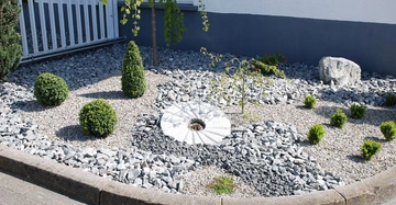 Ein gepflegter Steingarten mit verschiedenen Sträuchern und einem kleinen Baum liegt neben einem Gebäude mit grauer Fassade und Fenster flankiert von einem weißen Zaun und grauen Kieselsteinen.