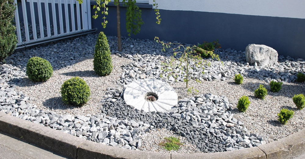 Ein gepflegter Steingarten mit verschiedenen Sträuchern und einem kleinen Baum liegt neben einem Gebäude mit grauer Fassade und Fenster flankiert von einem weißen Zaun und grauen Kieselsteinen.