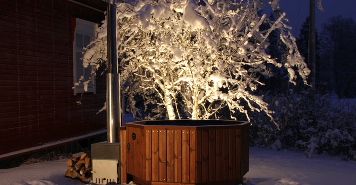 Galerie-Bild 2: Holzwanne im Freien steht vor einem verschneiten Baum und einem roten Gebäude im Hintergrund; das Ganze ist nachts und die Szene ist schneebedeckt und beleuchtet.