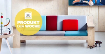 Holzsofa mit bunten Kissen steht in modernem Raum mit hellem Holz und Kunstwerken. Der gelbe Kreis enthält Text: "36 PRODUKT DER WOCHE". Ein Tisch links mit Kunstobjekt und Spielzeug rechts.